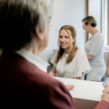 Drei Frauen sitzen an einem Empfangstresen, eine Person steht dahinter, helle Räume mit Fenstern